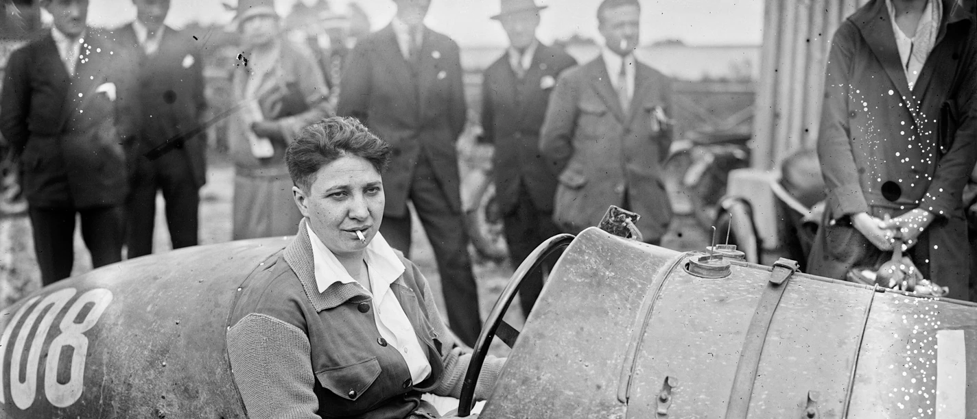 Violette Morris 1927 am Steuer eines Rennwagens, umringt von Zuschauenden