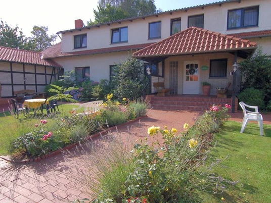 "Sonnenschein" Pension Haus Stork Ein zweigeschossiges Haus mit rotem Dach und Terrasse, umgeben von Rasenflächen und blühenden Beeten.
