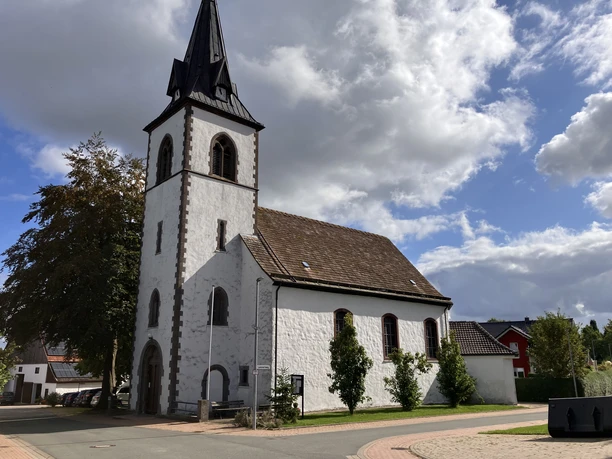 Weißer Kirchenbau mit Glockenturm im Ortskern, umgeben von Häusern und Bäumen.