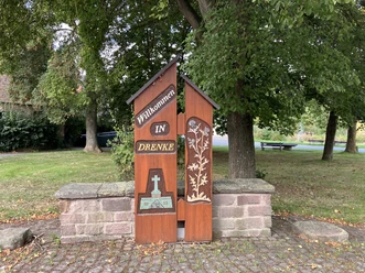 Hölzerne Ortstafel mit dem Schriftzug "Willkommen in Drenke" vor einer kleinen Steinmauer, umgeben von Bäumen.