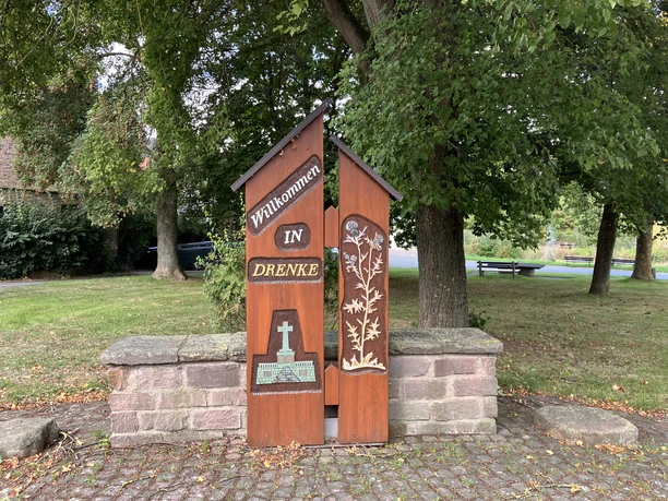 Hölzerne Ortstafel mit dem Schriftzug "Willkommen in Drenke" vor einer kleinen Steinmauer, umgeben von Bäumen.