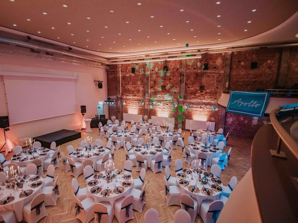 Festlich eingedeckter Apollo-Saal mit Rundtischen, Bühne und beleuchteter Backsteinwand.