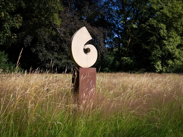 Helle Spiralskulptur von Gabriele Zutz auf rostigem Sockel in hoher Wiese vor Bäumen