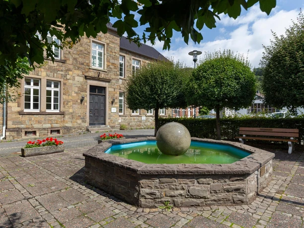 Kugelbrunnen auf gepflastertem Platz vor Natursteinhaus, umgeben von Bäumen und Bank