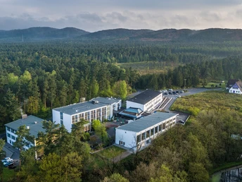 Luftbild Großes Gebäude inmitten eines grünen Waldgebiets, mit weitläufigen Parkflächen und Hügeln.