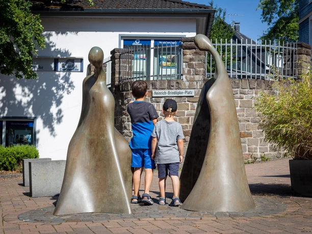 Zwei Kinder betrachten die Bronzeskulptur „Kommunikation“ auf einem Platz in Bielstein
