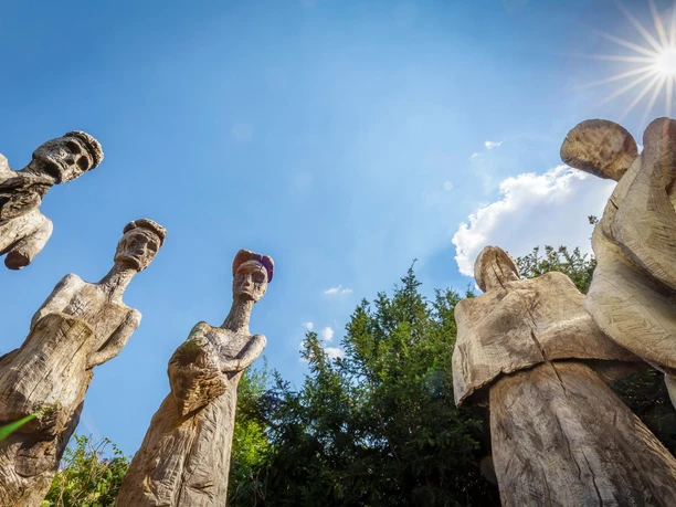 Holzskulpturen „Könige“ ragen unter blauem Himmel mit strahlender Sonne auf.
