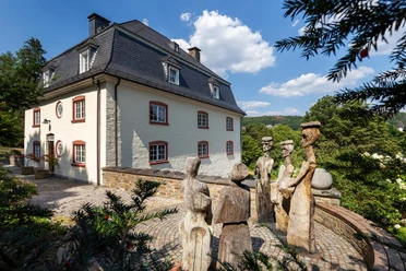 Holzskulptur „Könige“ vor hellem Haus mit Schieferdach in Wiehl-Bielstein
