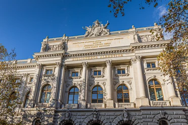 Historická fasáda Bibliotheca Albertina se sloupy, sochami a vysokými obloukovými okny je vidět na pozadí modré oblohy a je orámována stromy s pestrobarevným listím