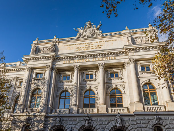 La façade historique de la Bibliotheca Albertina, avec ses colonnes, ses sculptures et ses hautes fenêtres cintrées, se détache sur fond de ciel bleu et est encadrée par des arbres aux feuilles colorées