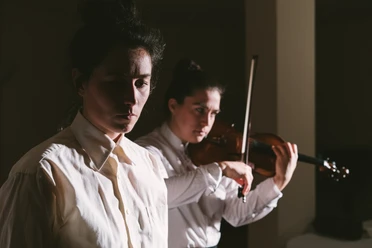 Two female musicians in white shirts, one holding a violin, bathed in dramatic light