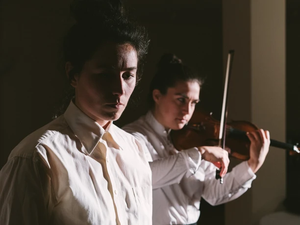 Two female musicians in white shirts, one holding a violin, bathed in dramatic light