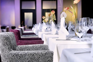 Bequeme Sessel stehen vor gedeckten Tischen mit weißer Tischdecke. Die Wand im Hintergrund ist Violet und passt ins Ensemble.