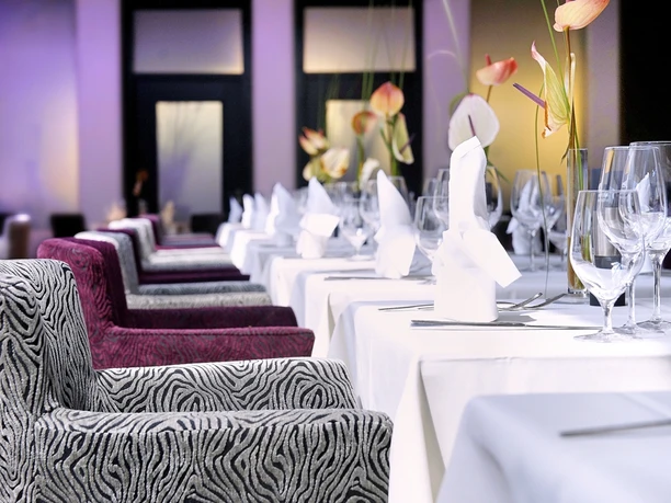 Bequeme Sessel stehen vor gedeckten Tischen mit weißer Tischdecke. Die Wand im Hintergrund ist Violet und passt ins Ensemble.