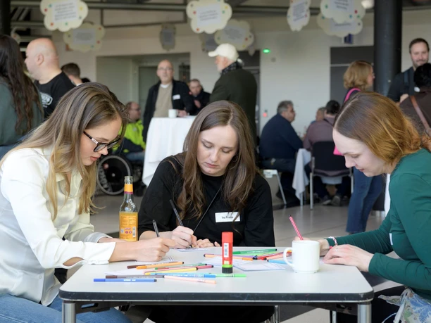 Drei Frauen schreiben bei einer Inklusionskonferenz an einem Tisch, im Hintergrund Gäste.