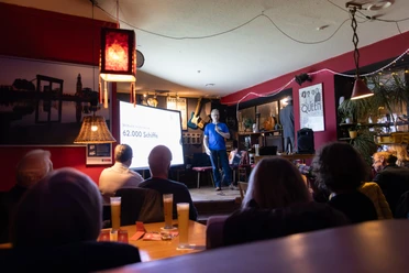 Vortrag im Café de Fiets mit Publikum, Leinwand und Sprecher auf kleiner Bühne.