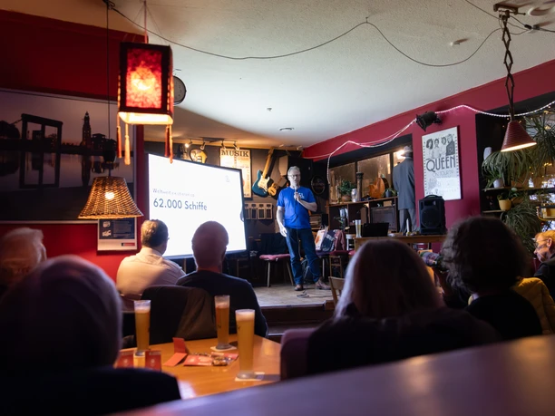 01_Fiets-10.jpg Vortrag im Café de Fiets mit Publikum, Leinwand und Sprecher auf kleiner Bühne.