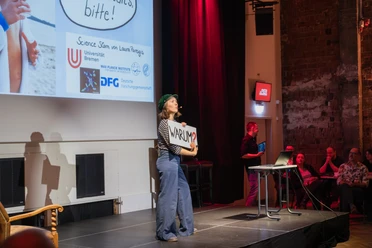 Referentin beim Science Slam auf der Bühne, mit „Warum?“-Schild vor Publikum.