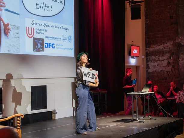 SL-82.jpg Referentin beim Science Slam auf der Bühne, mit „Warum?“-Schild vor Publikum.