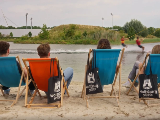 Wakepark.jpg Vier Menschen sitzen gemütlich in Klappstühlen am Sand. Zwei fahren auf dem Wasser Wakeboard.