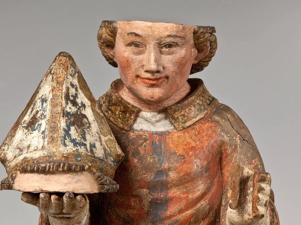 Bemalte Holzfigur des heiligen Dionysius mit Mitra und Buch vor grauem Hintergrund
