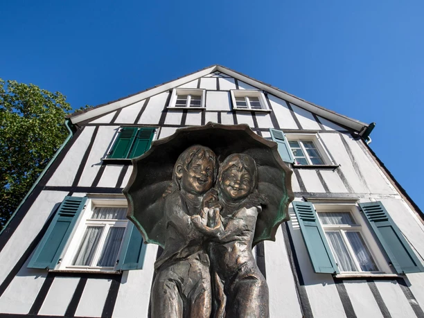 Kinder unterm Regenschirm Bronzeskulptur zweier Kinder unter einem Schirm vor einem Fachwerkhaus in Waldbröl