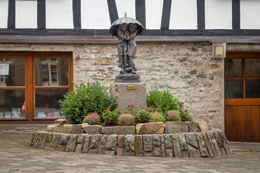 Bronzeskulptur zweier Kinder unter Regenschirm auf bepflanztem Sockel vor Fachwerkhaus