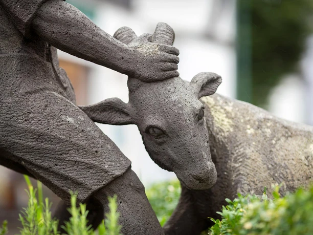 Steinfigur am Ziegenbrunnen: Eine Hand hält den Kopf einer Ziege zwischen grünen Pflanzen