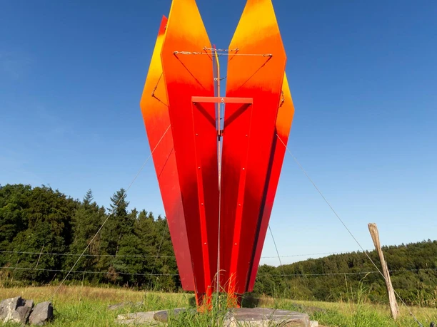 Rote Skulptur „himmelwärts“ auf einer Wiese vor blauem Himmel