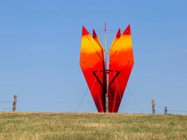 Rot-orange Skulptur „himmelwärts“ auf einer Wiese vor blauem Himmel