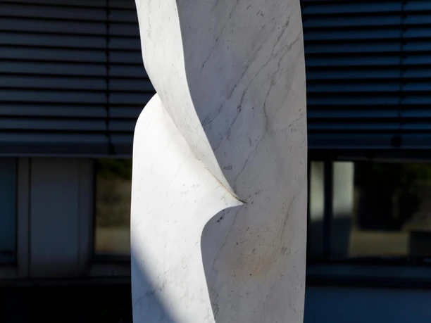 Weiße Marmorskulptur „Kraft II“ auf Sockel vor moderner Fassade