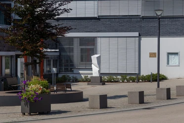 Weiße Steinskulptur „Kraft II“ auf einem Platz vor moderner Fassade in Nümbrecht