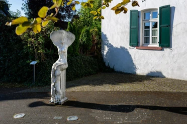 Skulptur „Erinnerung an einen Baum“ vor weißem Haus mit grünen Fensterläden