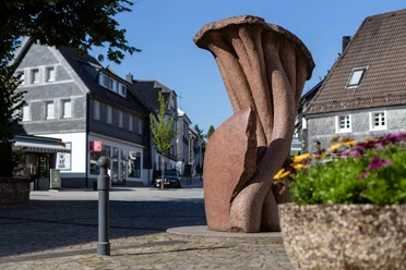 Rötliche Steinskulptur „Vergänglichkeit“ von Georg Weber in einer Straße in Nümbrecht