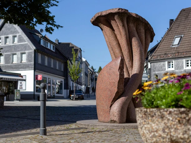 Rötliche Steinskulptur „Vergänglichkeit“ von Georg Weber in einer Straße in Nümbrecht