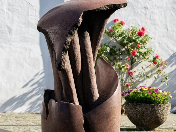 Rostbraune Skulptur „Vergänglichkeit“ vor weißer Wand mit blühenden Rosen.