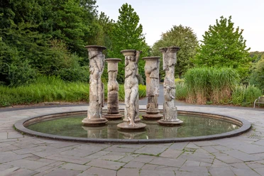 Säulenbrunnen von Michael Schwarze mit Figurenreliefs in rundem Becken, umgeben von Grün