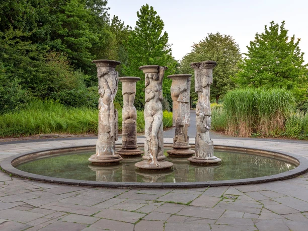 Säulenbrunnen von Michael Schwarze mit Figurenreliefs in rundem Becken, umgeben von Grün