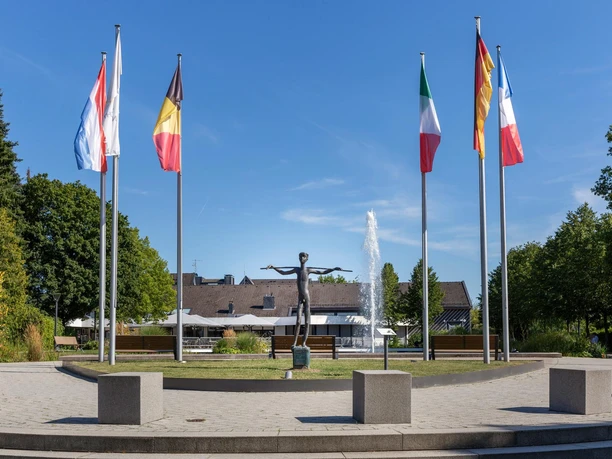Bronzeskulptur „Knabe mit Stab“ zwischen Fahnen und Brunnen in Nümbrecht