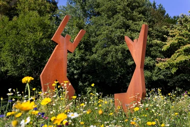 Rostrote Skulptur „Freude“ von Rosa Gilissen-Vanmarcke in einer bunten Blumenwiese