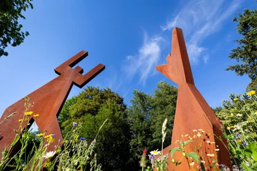 Rostrote Stahlskulptur „Freude“ zwischen Wildblumen vor blauem Himmel in Nümbrecht