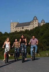 Vier Personen fahren auf Fahrrädern vor einer majestätischen Burg auf einem Hügel entlang.