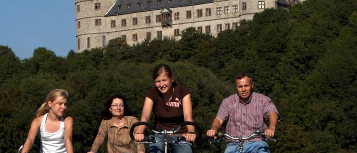 Vier Personen fahren auf Fahrrädern vor einer majestätischen Burg auf einem Hügel entlang.