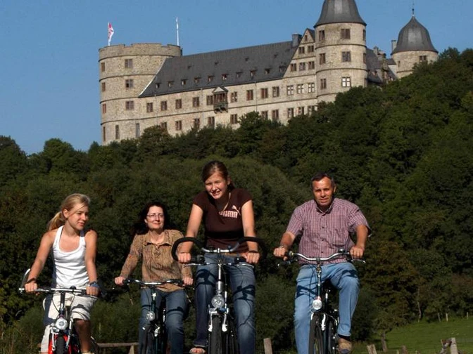 Vier Personen fahren auf Fahrrädern vor einer majestätischen Burg auf einem Hügel entlang.