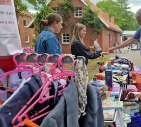flohmarktAKSR.jpg Flohmarktstand mit Kleidung, Spielzeug und Besuchern vor rotem Backsteinhaus.