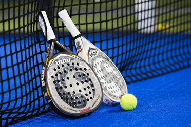 Padel-Am-Meer.jpg Zwei Padelschläger und ein Ball liegen am Netz auf blauem Sportbelag.