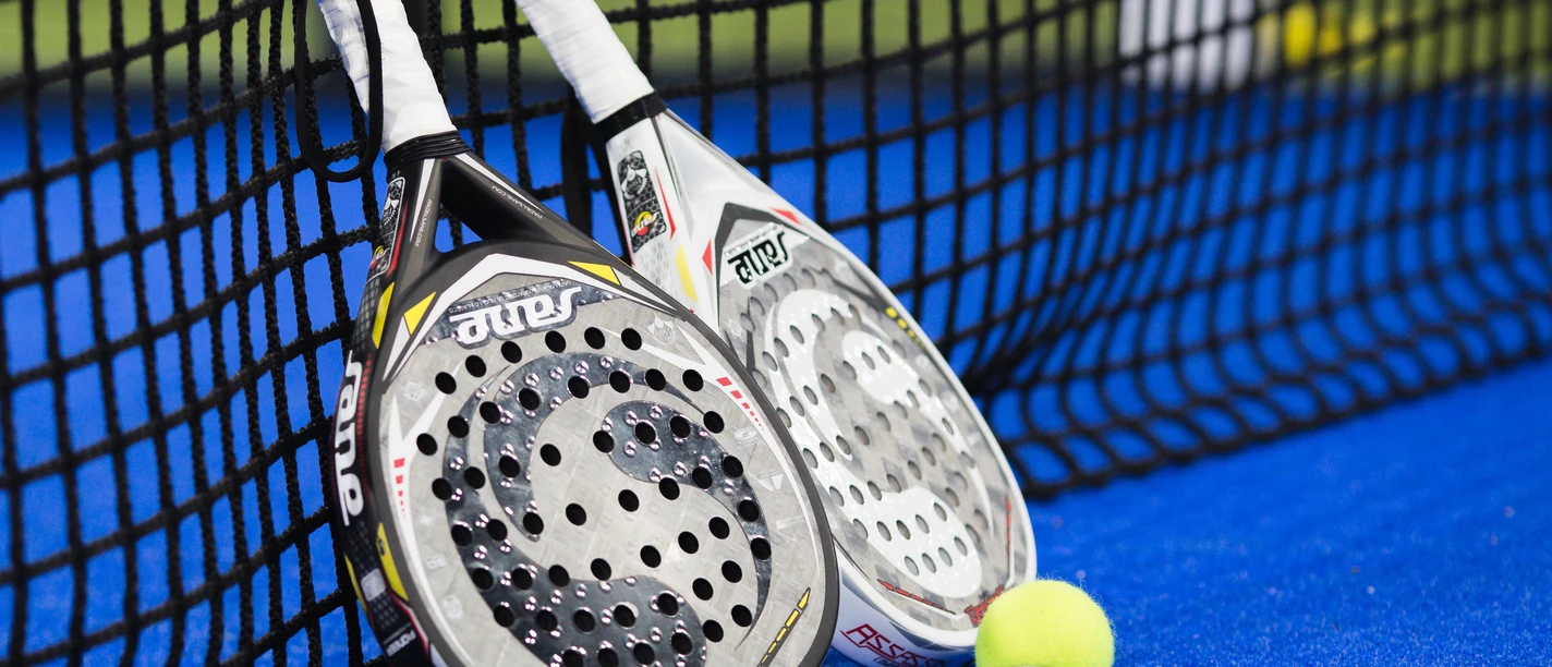 Zwei Padelschläger und ein Ball liegen am Netz auf blauem Sportbelag.