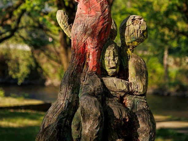 Holzskulptur mit roten und grünen Figuren vor Bäumen im Sinneswald
