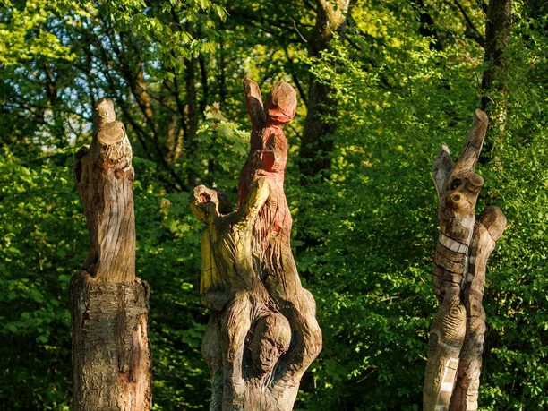 Drei Holzskulpturen mit menschlichen Figuren im grünen Sinneswald bei Sonnenlicht
