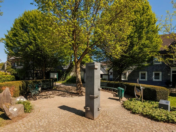 Steinernes Denkmal auf dem Friedrich-Ueberweg-Platz, umgeben von Bänken und grünen Bäumen